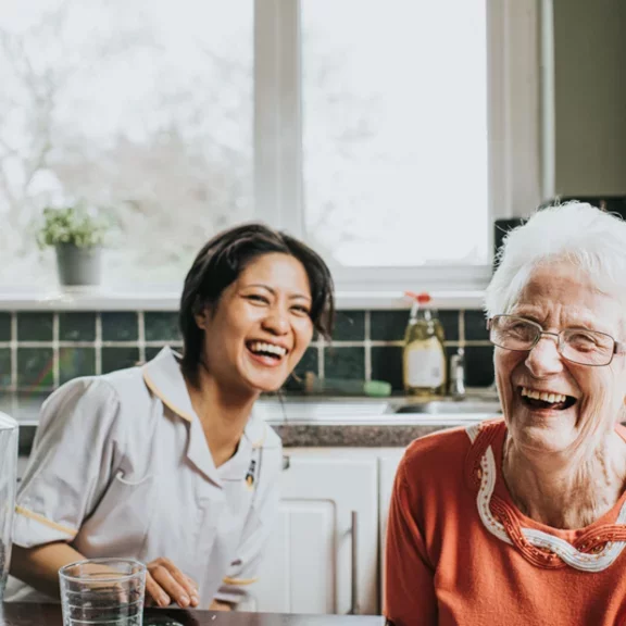 An elderly woman laughs beside a friendly young care assistant